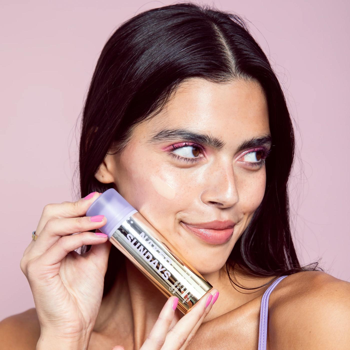 Woman holding Naked Sundays CabanaClear illuminating water gel serum SPF50 bottle near her cheek