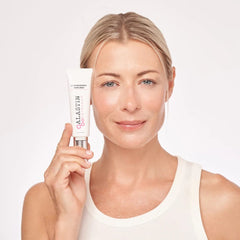 Woman with glowing skin holding ALASTIN Ultra Nourishing Moisturizer tube beside her face