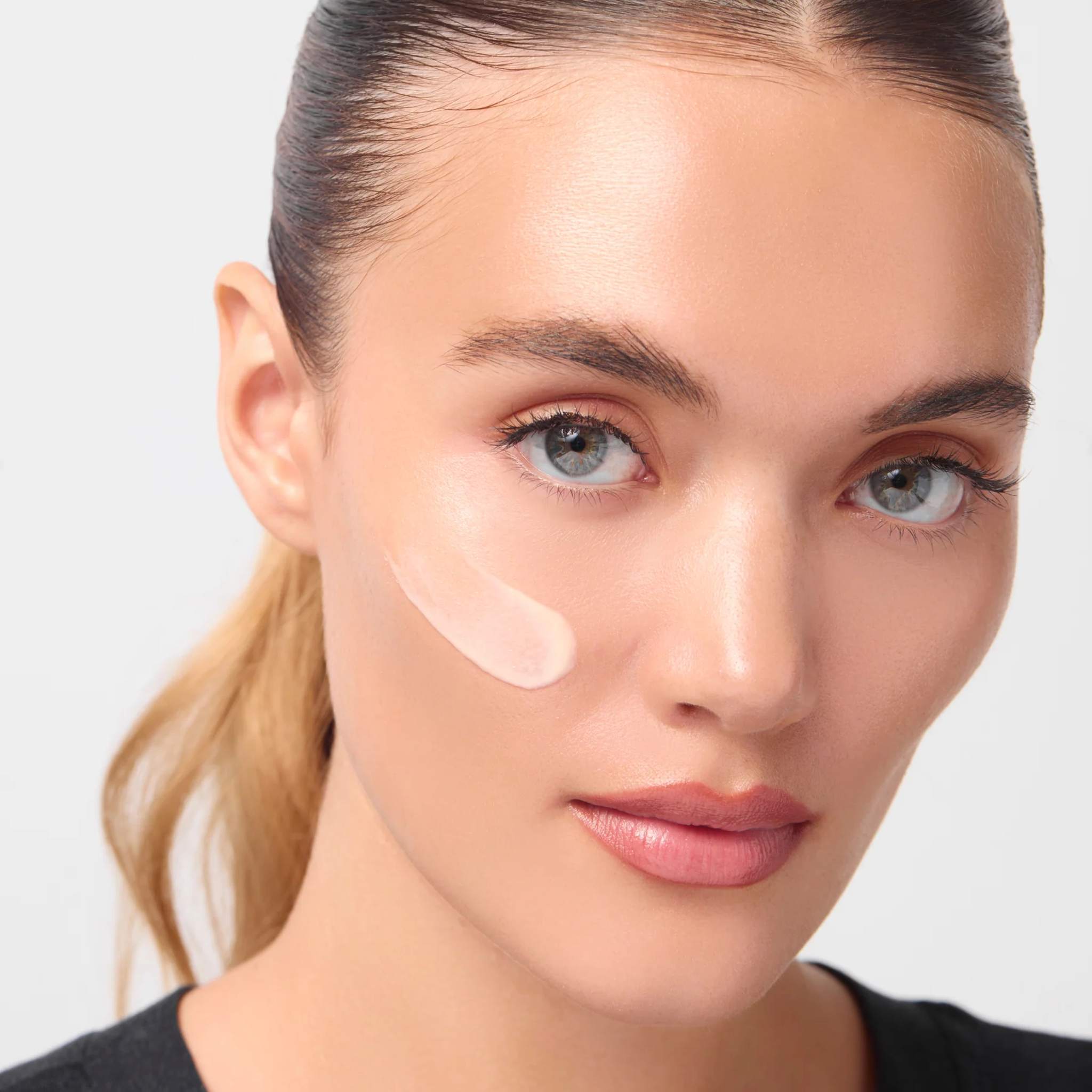 Woman with slicked back ponytail wearing a swipe of peach firming daily treatment cream on her cheek