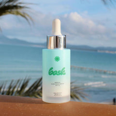 Bask SPF 30 mineral sunscreen serum dropper bottle on ledge with tropical ocean and palm tree background