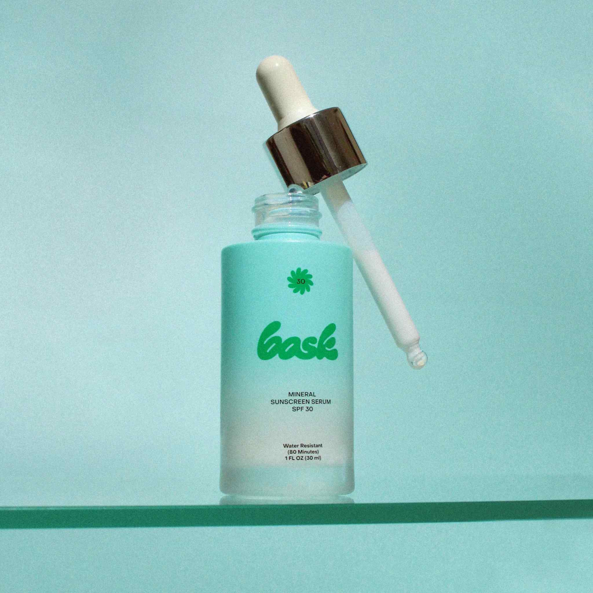 Bask SPF 30 mineral sunscreen serum open frosted bottle with dropper leaning against it on teal surface