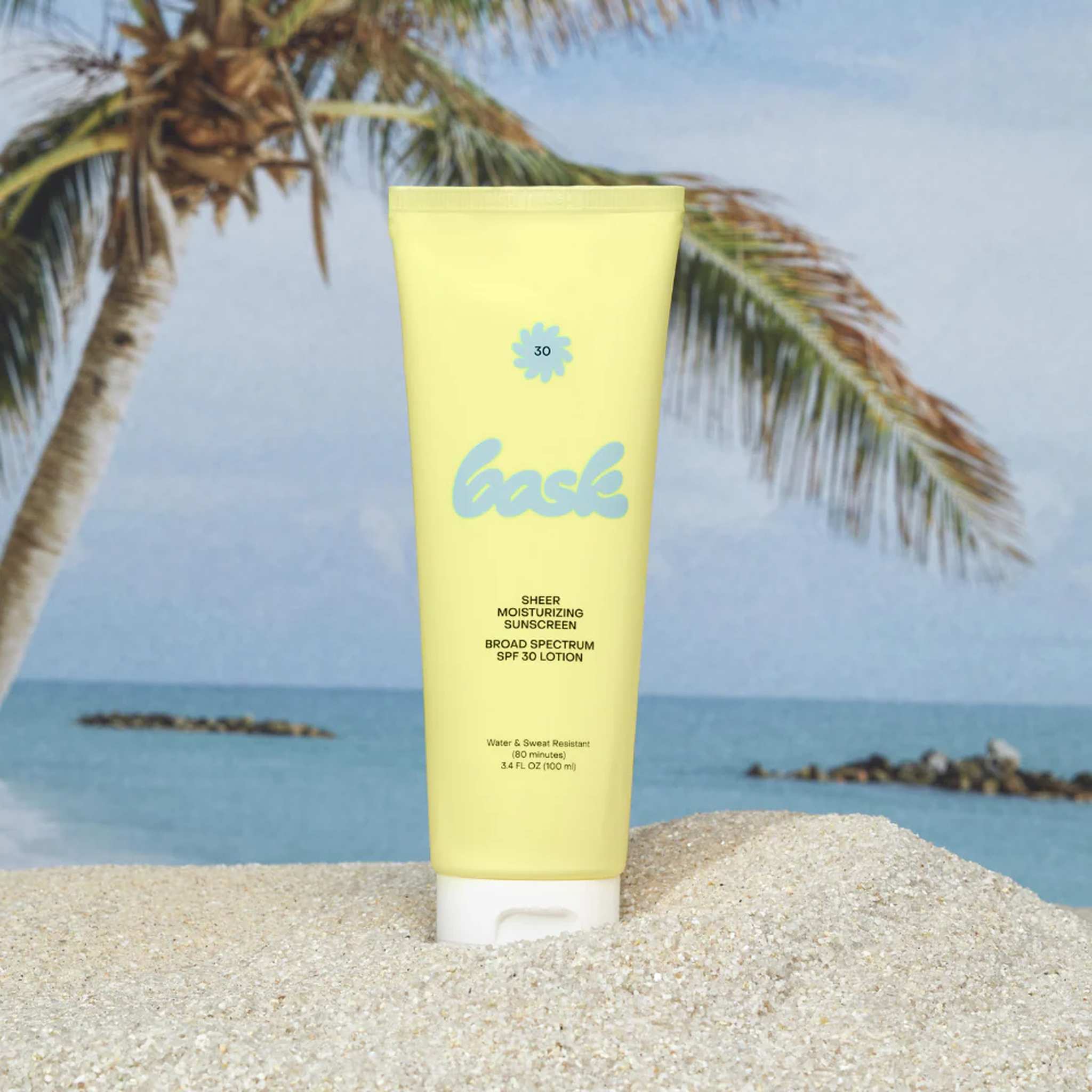 Bask SPF 30 sheer moisturizing sunscreen lotion tube standing in sand with tropical palm tree and ocean backdrop