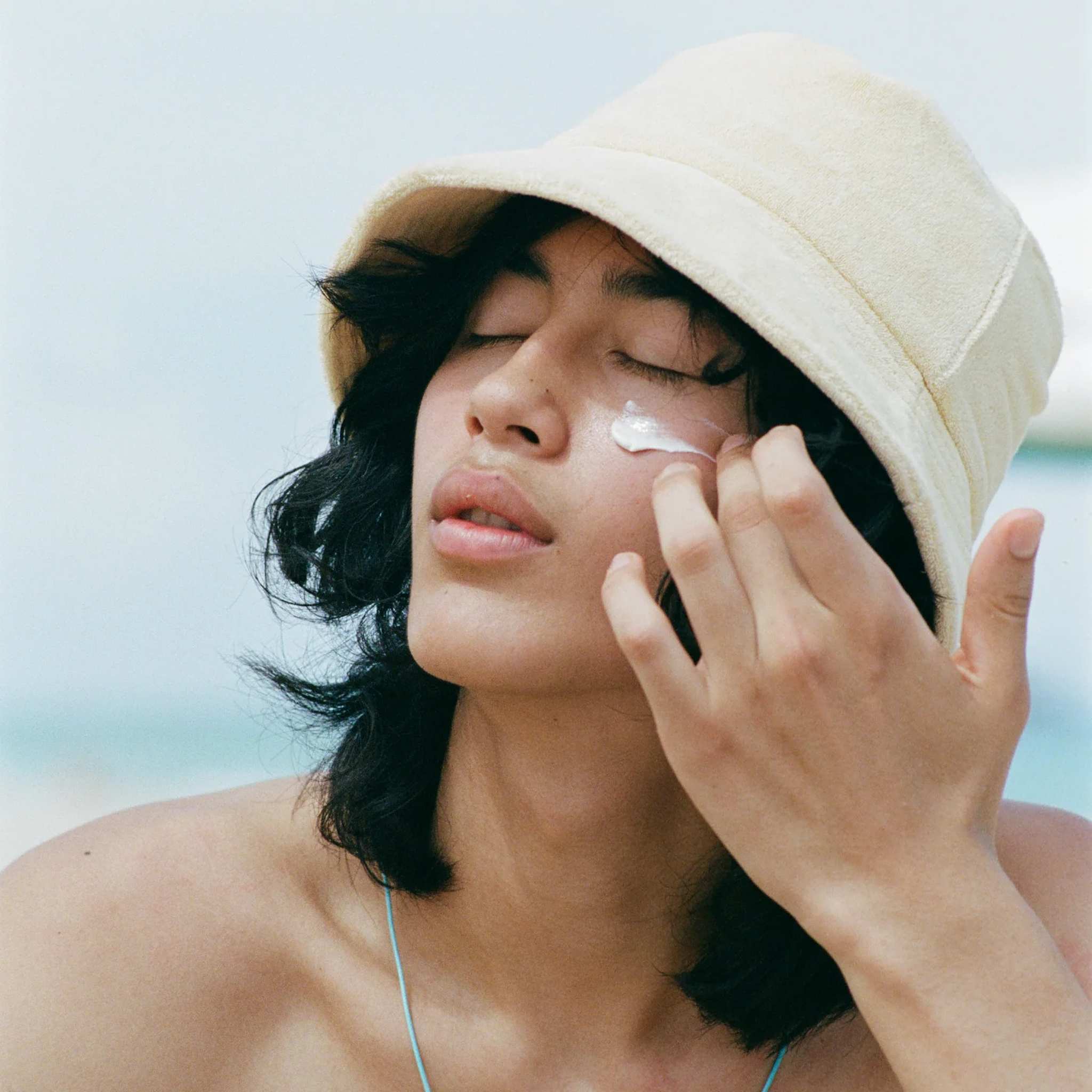 Model in bucket hat applying Bask SPF 30 sheer moisturizing sunscreen lotion to face at the beach