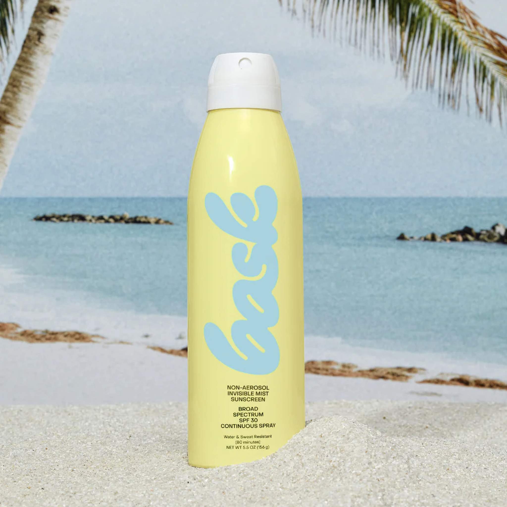 Bask SPF 30 invisible mist sunscreen spray standing on sandy beach with tropical ocean and palm tree backdrop