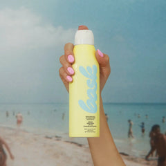 Hand with pink nails holding Bask SPF 30 non-aerosol sunscreen spray above sunny beach ocean scene