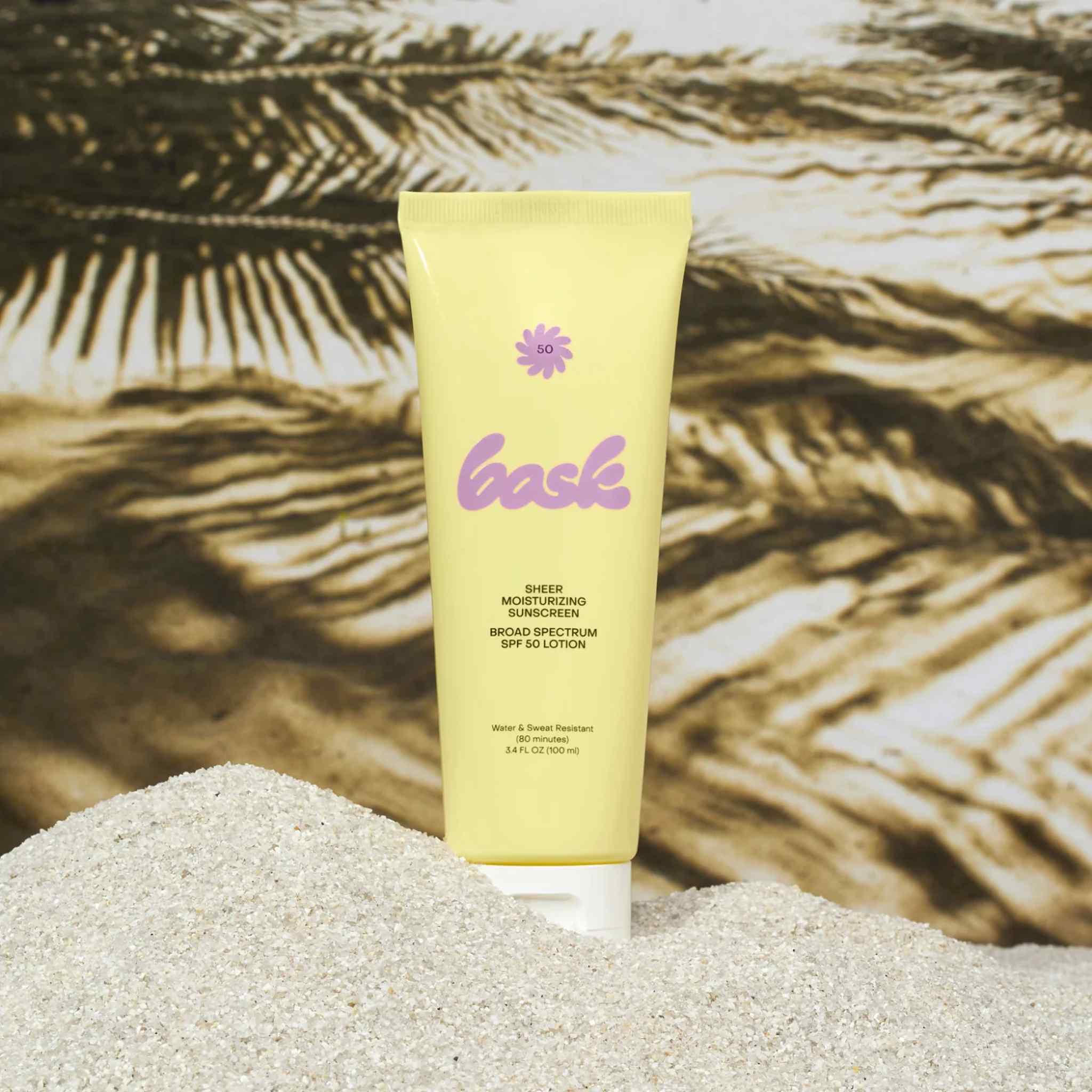 Bask SPF 50 sheer moisturizing sunscreen lotion tube standing in sand with tropical palm tree shadow background