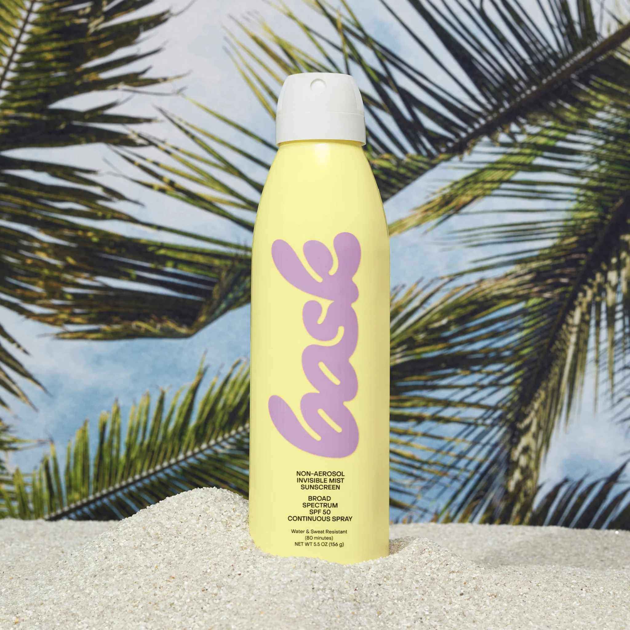 Bask SPF 50 invisible mist sunscreen spray standing in sand with tropical palm tree backdrop