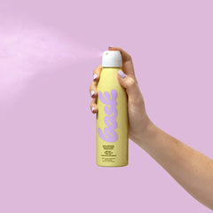 Hand holding Bask SPF 50 invisible mist sunscreen spray bottle against lavender purple background