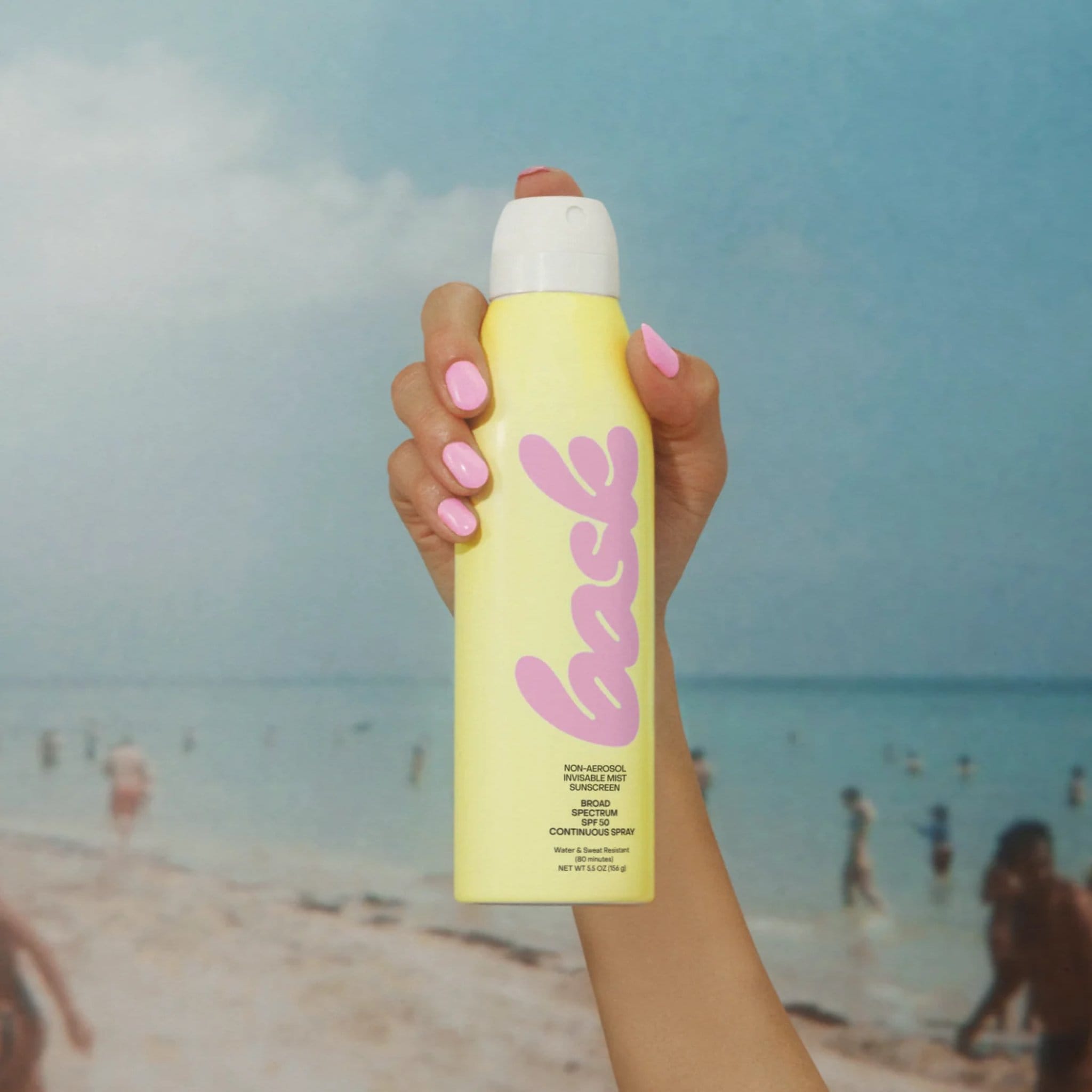 Hand holding Bask SPF 50 non-aerosol sunscreen spray above beach ocean scene