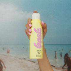 Hand holding Bask SPF 50 non-aerosol sunscreen spray above beach ocean scene
