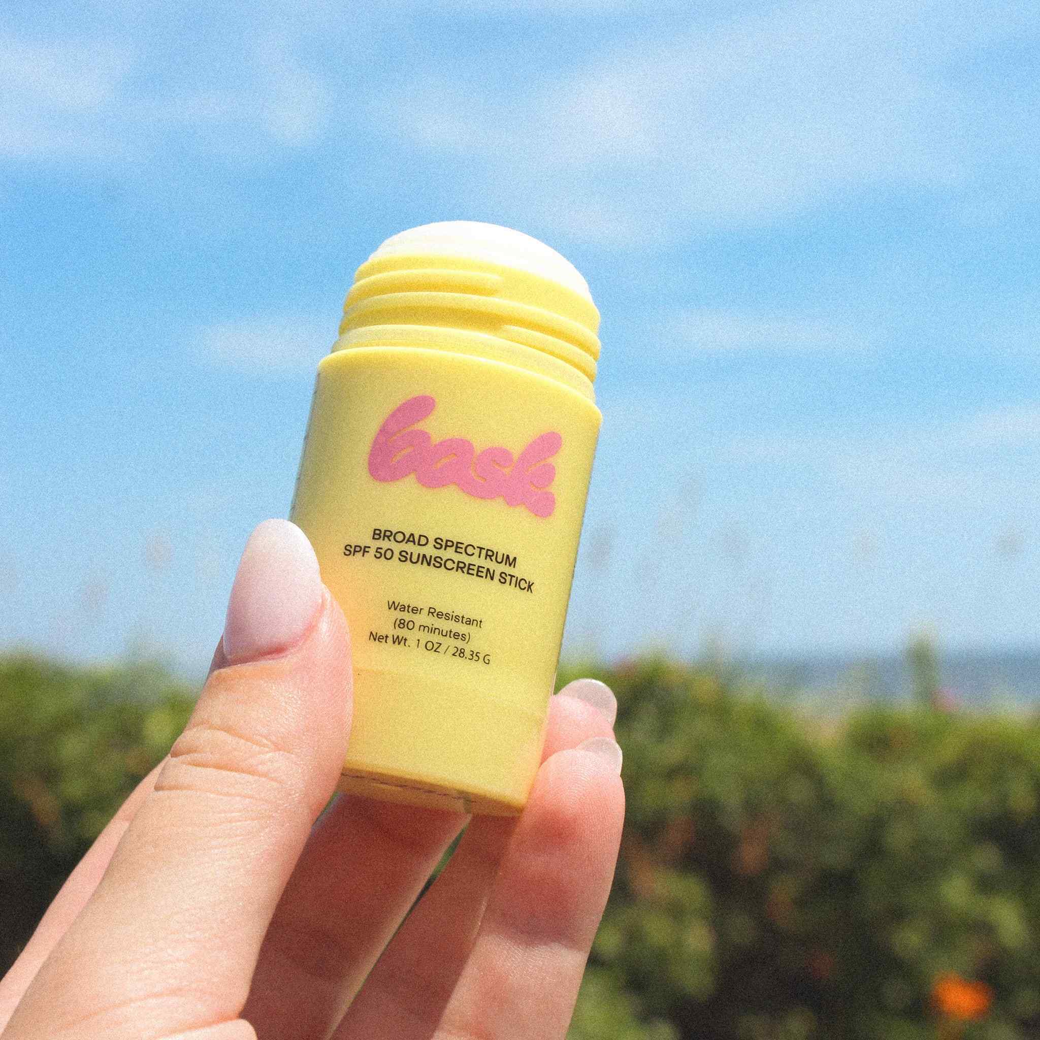Hand holding Bask SPF 50 sunscreen stick against blue sky and greenery outdoor background