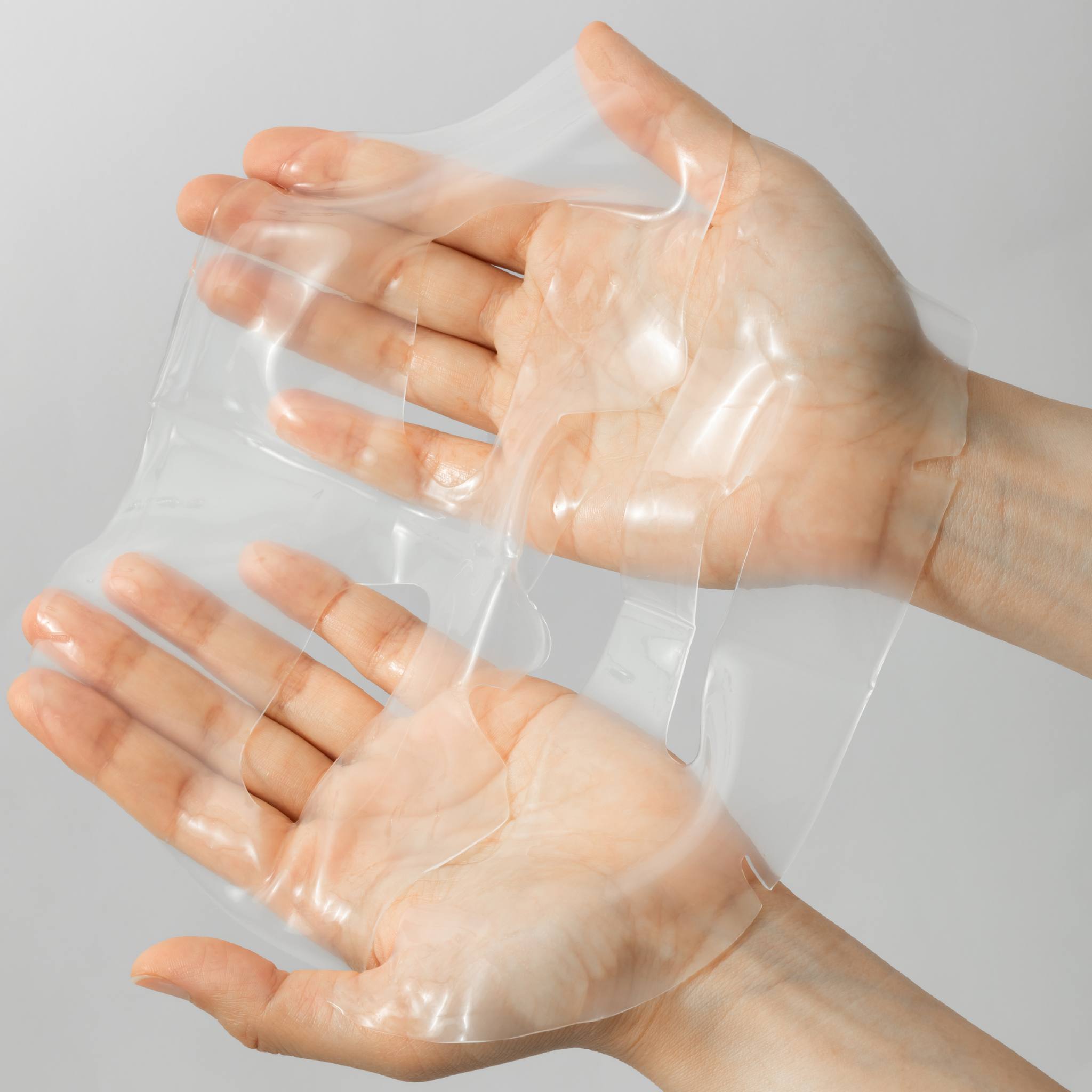 Hands holding translucent BIOEFFECT Imprinting Hydrogel Mask sheet showing texture and flexibility of hyaluronic acid face mask
