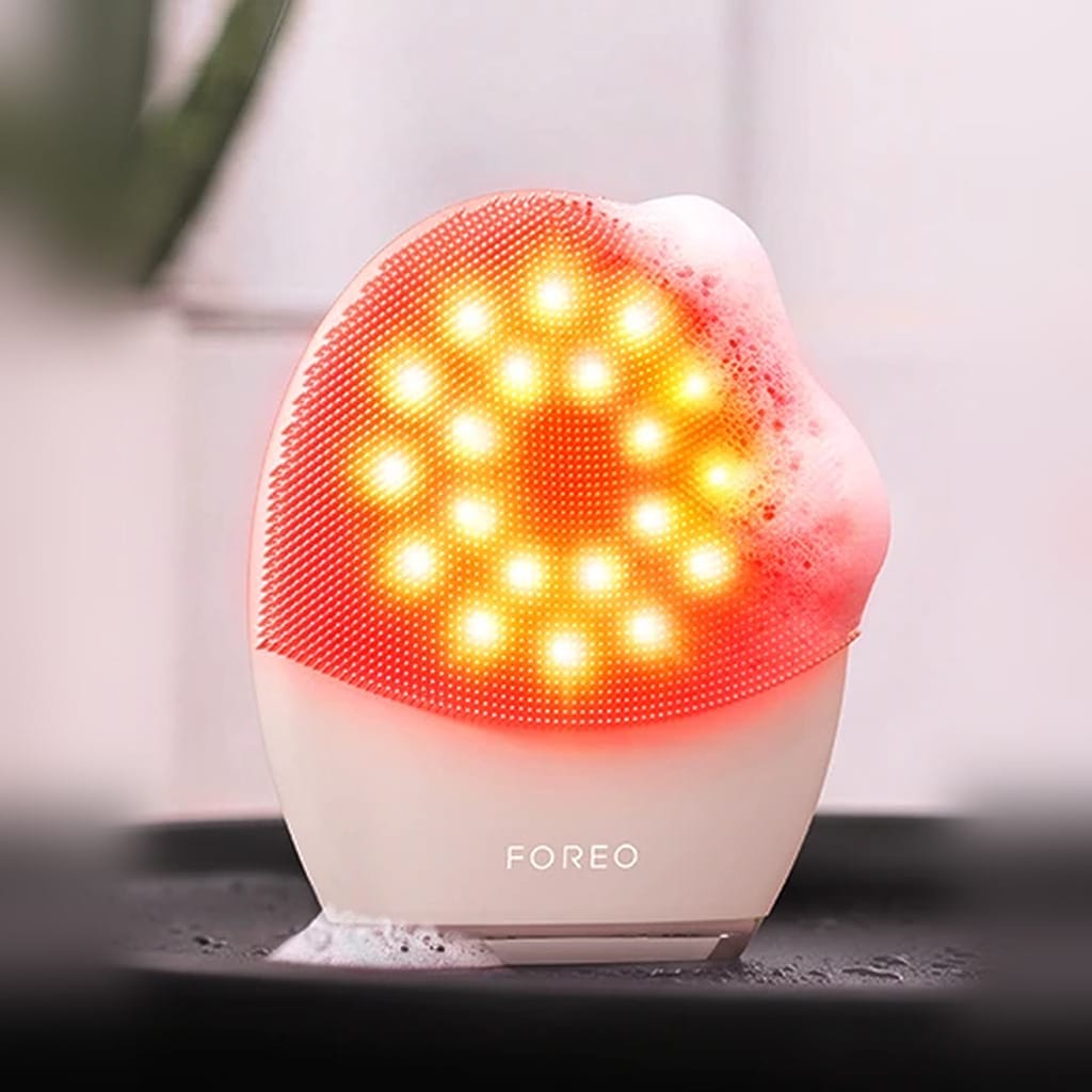FOREO LUNA 5 LED silicone face brush glowing with red LED light and cleanser foam on a bathroom tray