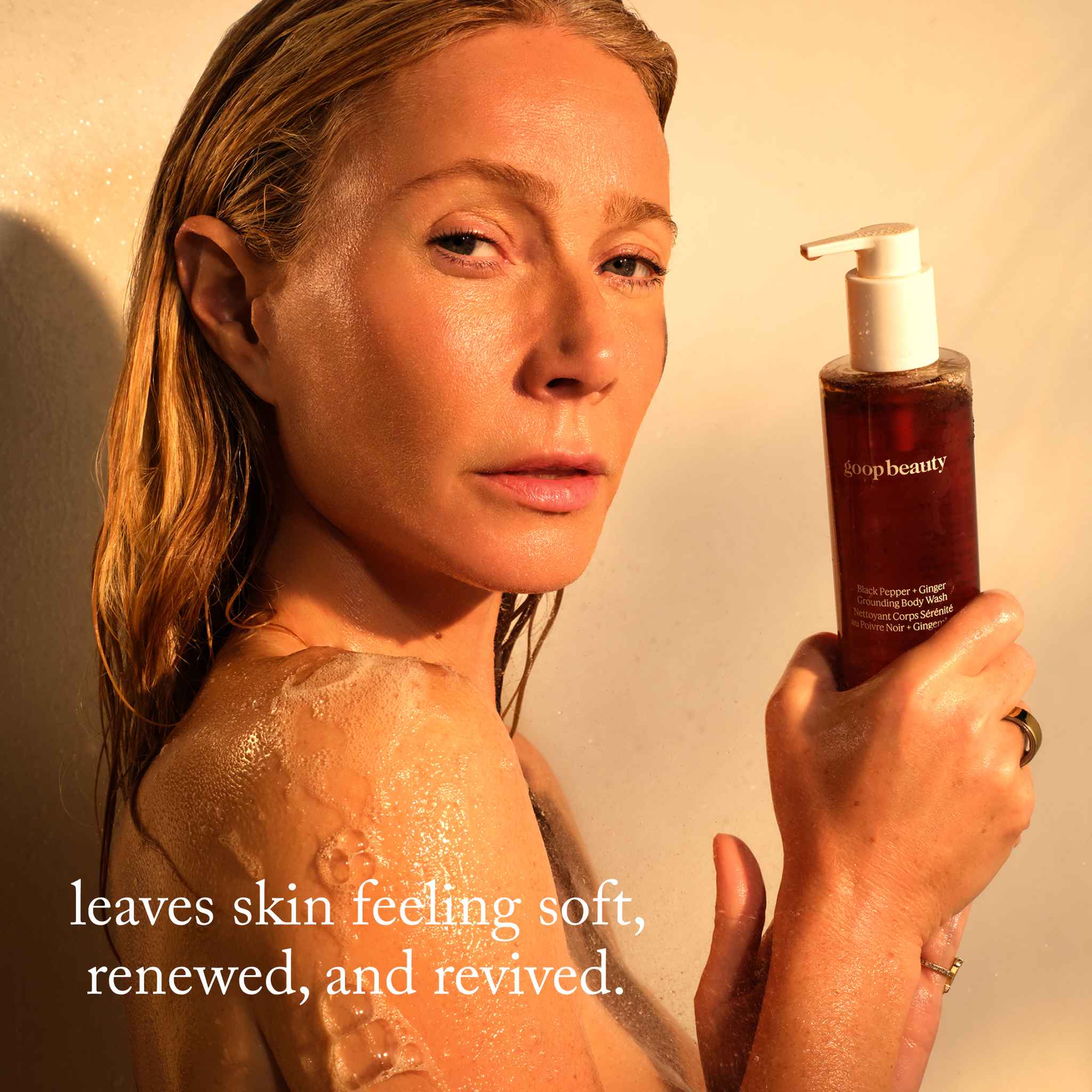 lifestyle-model: Woman holding goop beauty Black Pepper Ginger body wash bottle with lather on her shoulder in warm light