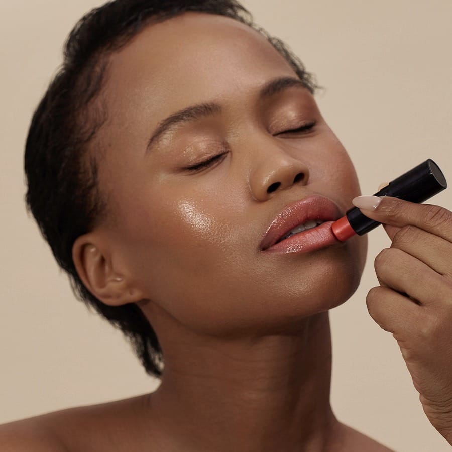 Woman with short curls applying Henné Organics Luxury Lip Tint in Coral for a glossy coral lip 2
