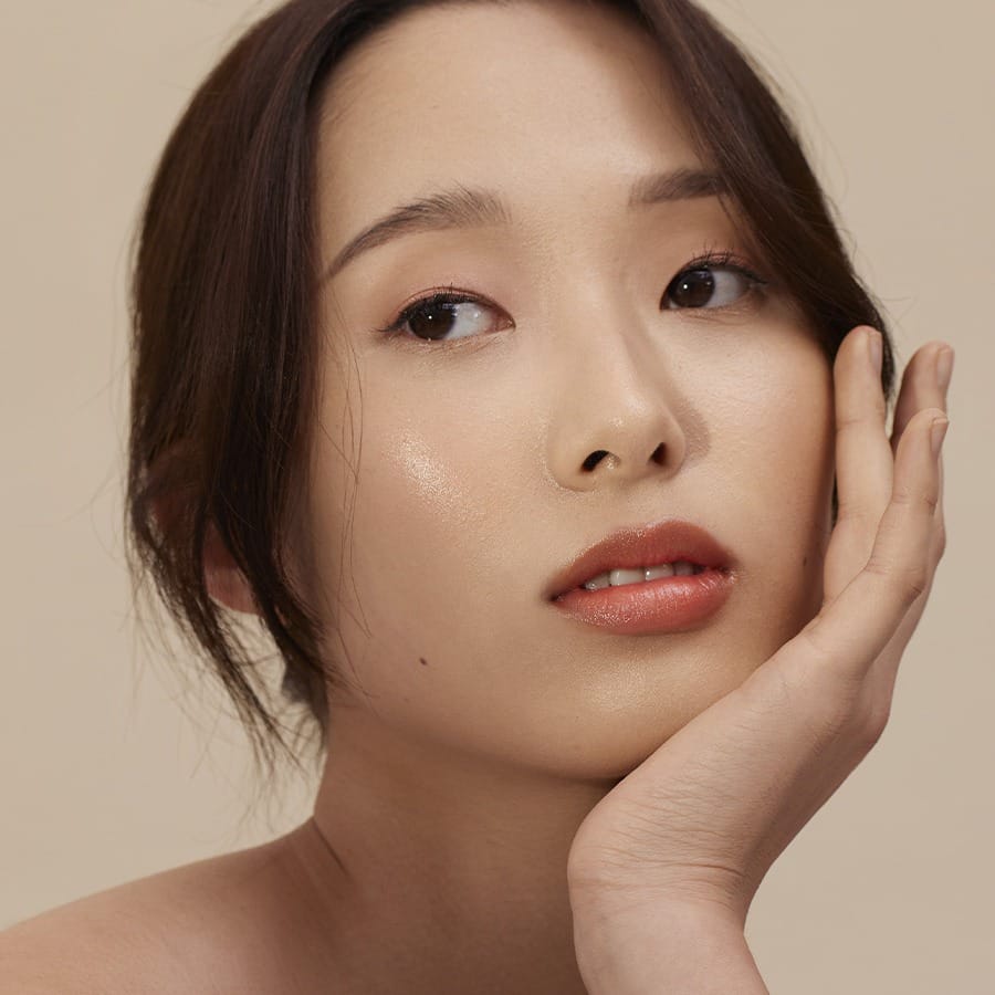 Model with hand on cheek wearing Henné Organics Luxury Lip Tint in Coral for a sheer coral lip 5