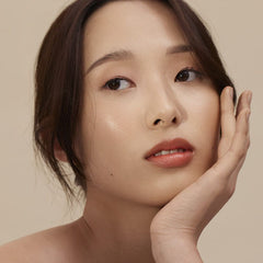 Model with hand on cheek wearing Henné Organics Luxury Lip Tint in Coral for a sheer coral lip 5