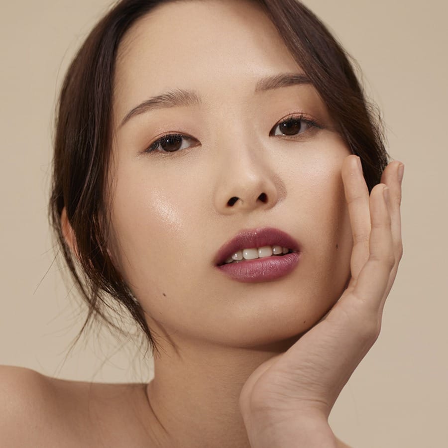 Model with hand on cheek wearing Henné Organics Luxury Lip Tint in Muse for a glossy plum lip 3