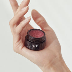 Hand holding an open jar of Henné Organics Lip Exfoliator filled with berry pink sugar scrub