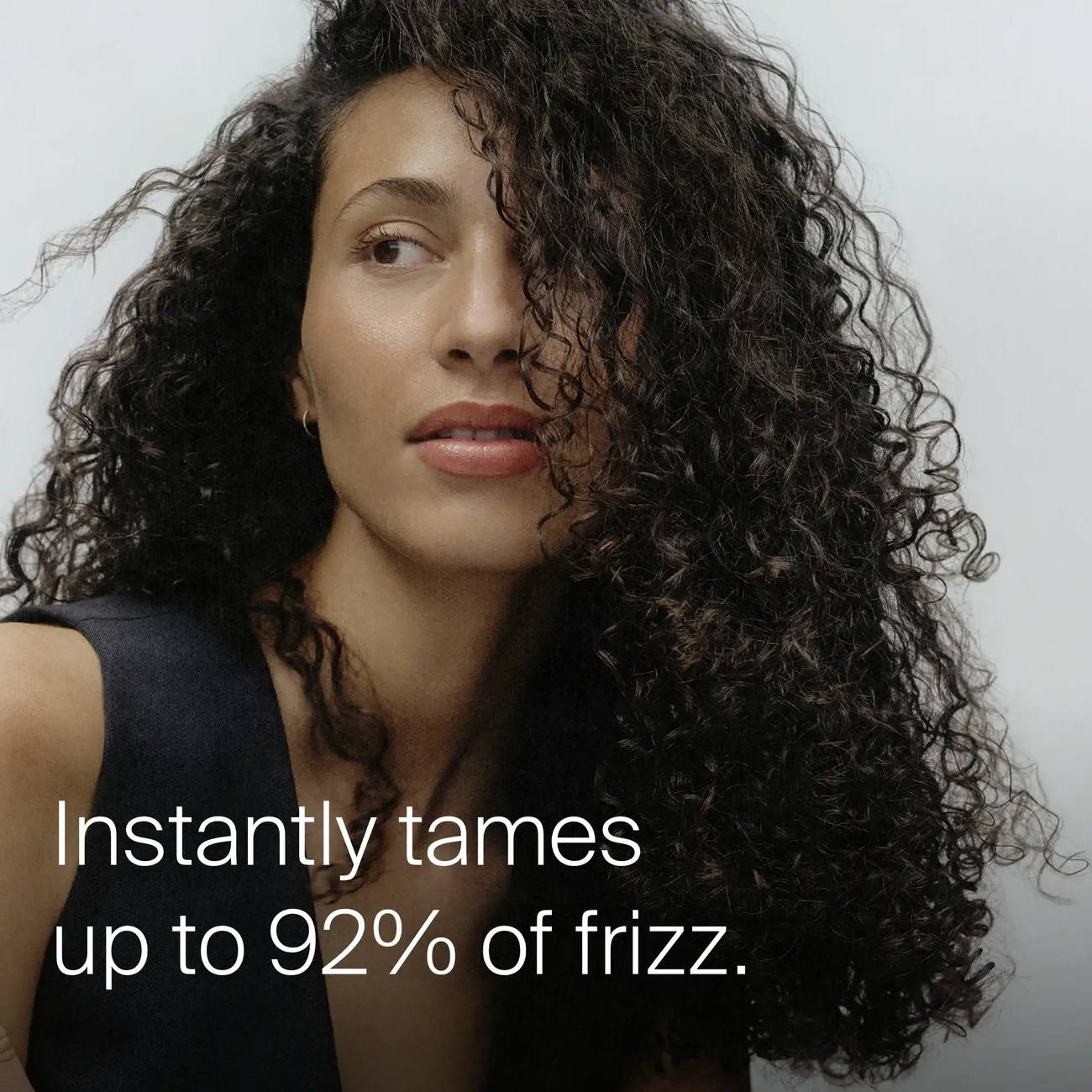 Woman with long dark curls showing Living Proof No Frizz Instant De-Frizzer taming up to 92 percent of frizz