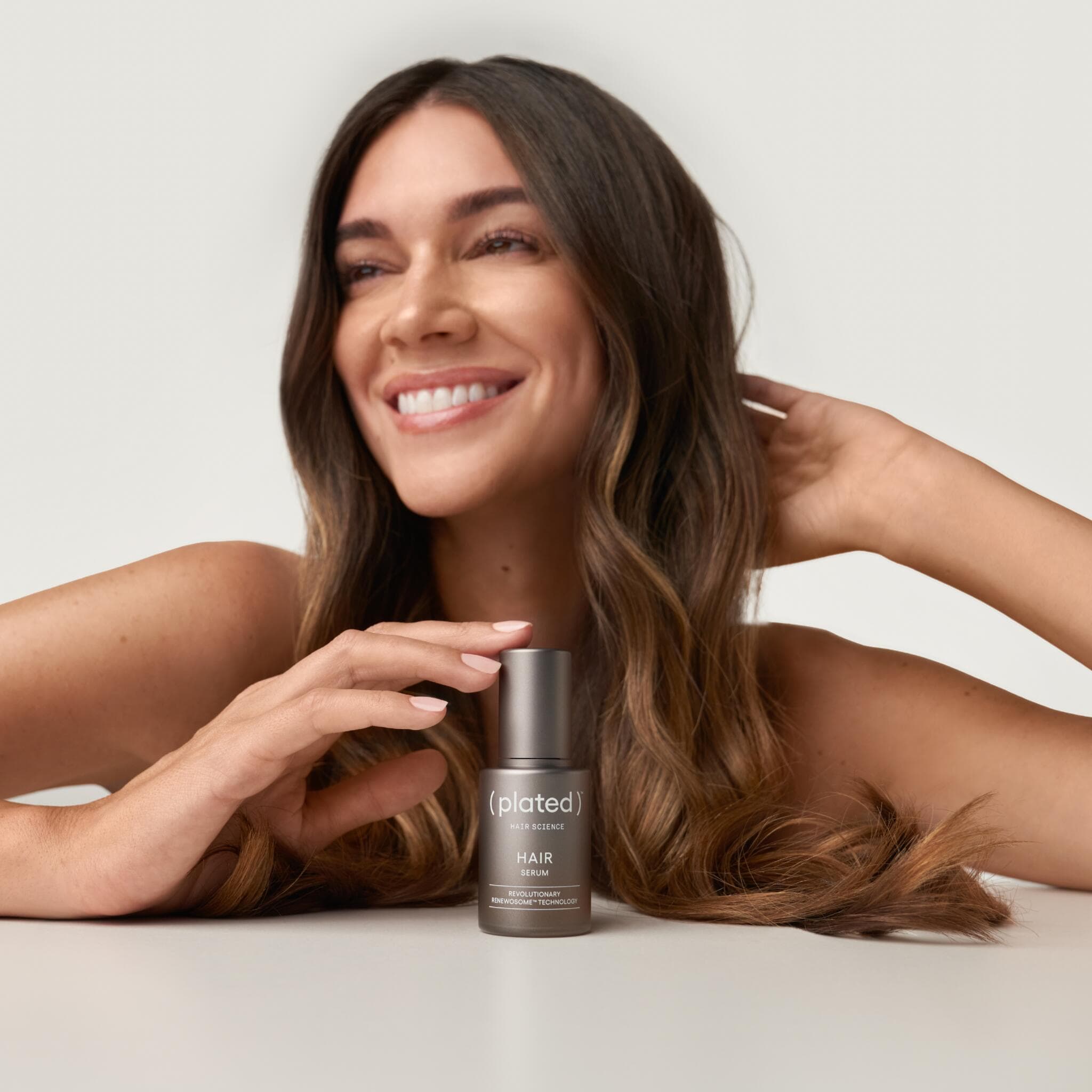 Smiling woman with wavy hair holding Plated Hair Serum bottle