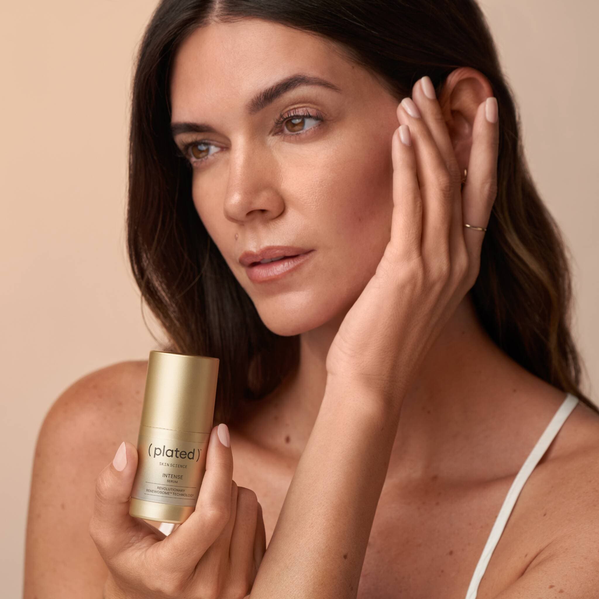 Woman holding Plated Intense Serum bottle while touching her face