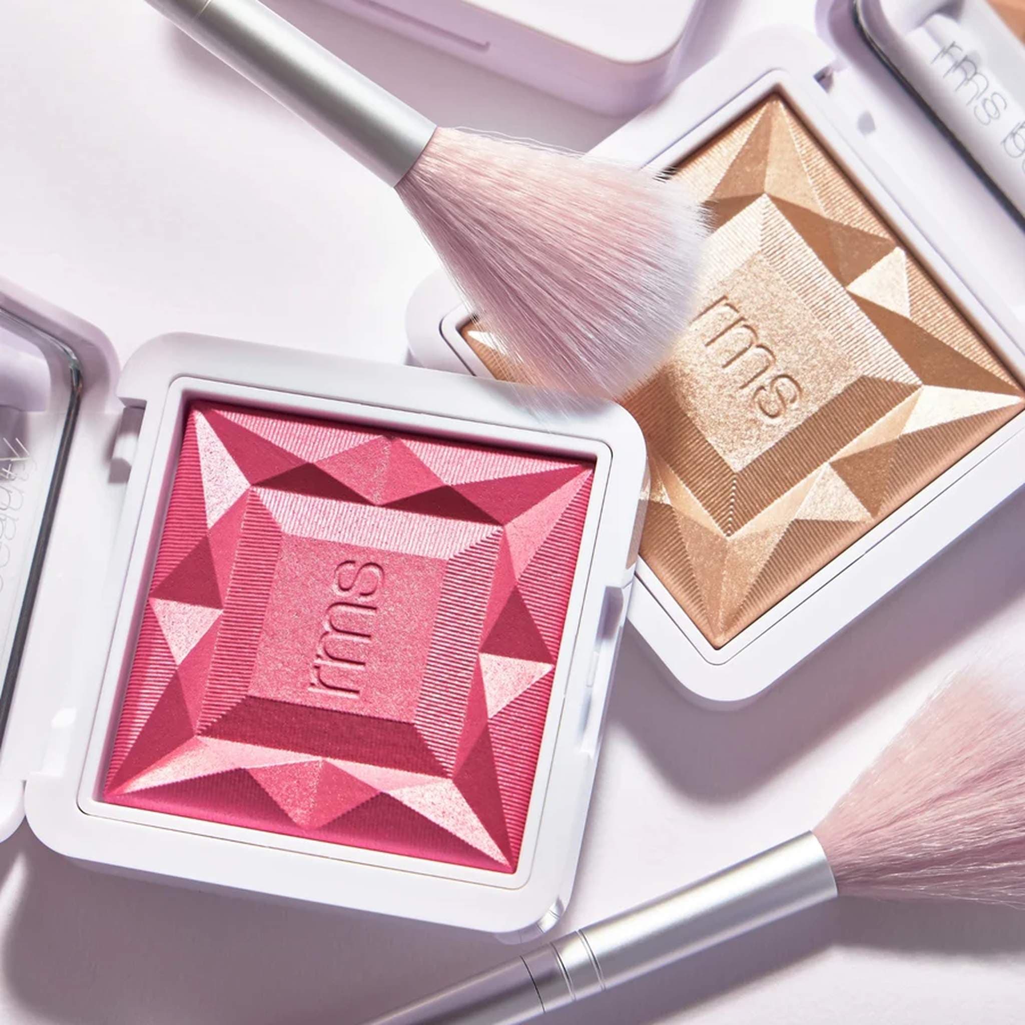 Fan brush resting on RMS Beauty luminizer and blush compacts with faceted texture