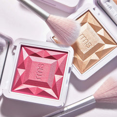 Fan brush resting on RMS Beauty luminizer and blush compacts with faceted texture
