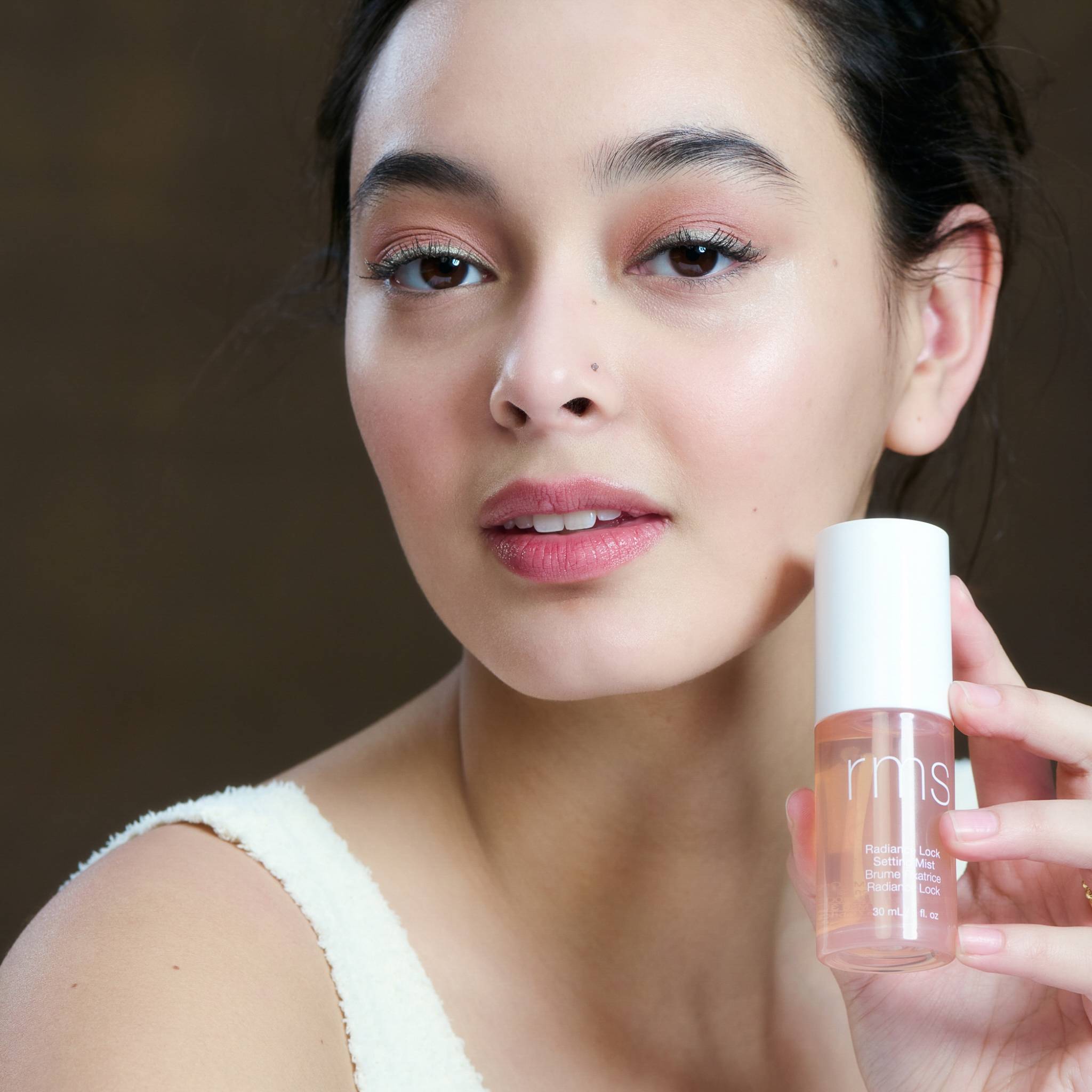 Model holding the 30ml travel size Radiance Lock Setting Mist near her cheek