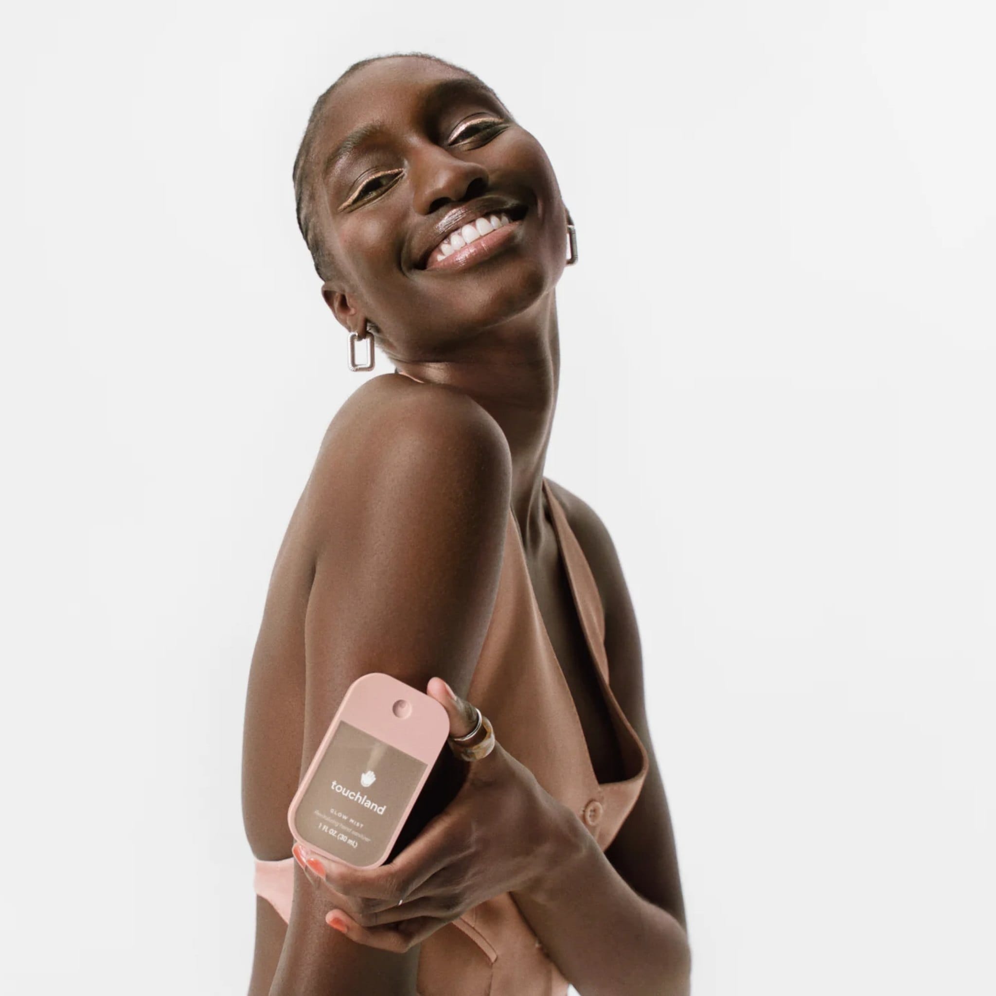 Smiling woman in a blush halter top holding Touchland Glow Mist Rosewater hand sanitizer against her arm