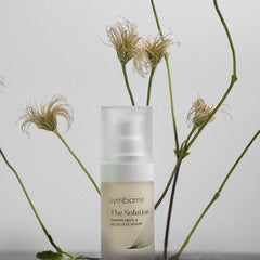 Symbiome The Solution Firming Neck and Decollete Serum bottle surrounded by wispy dried botanical stems on gray surface