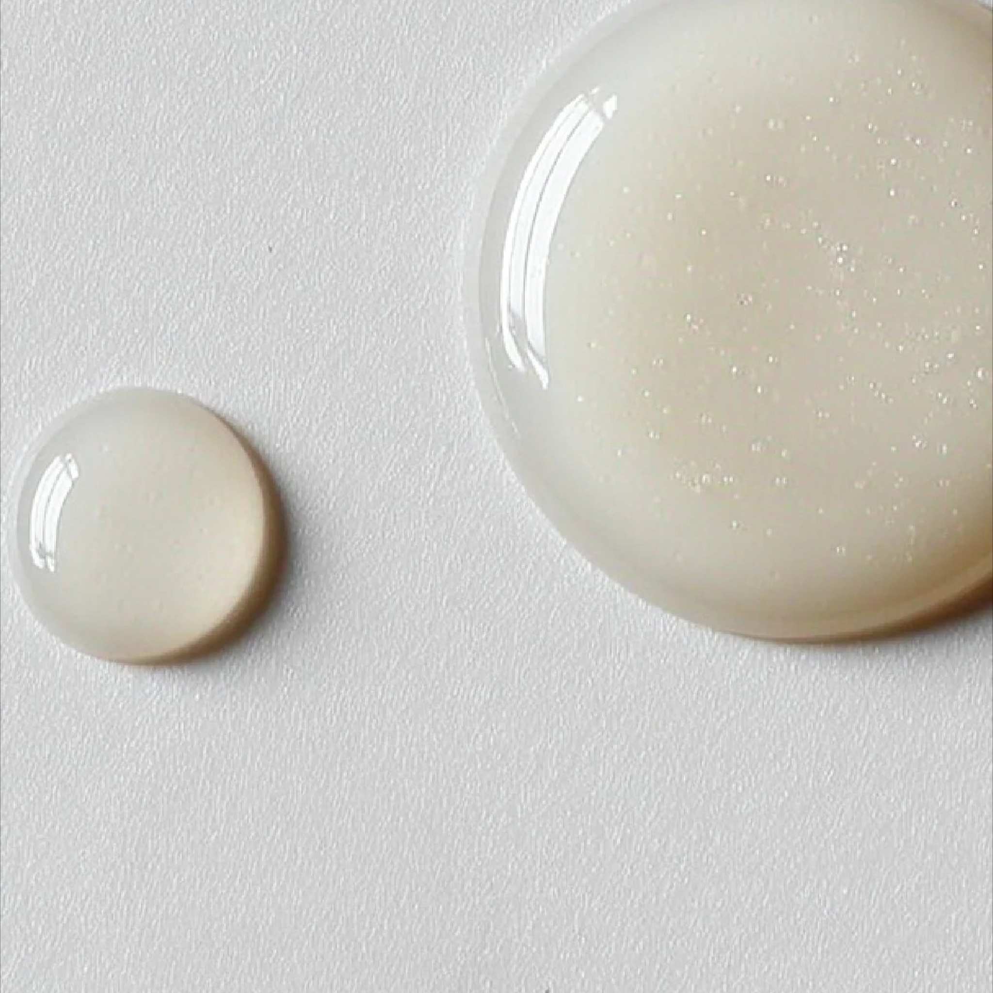 Two pearlescent serum droplets on white surface showing the lightweight gel texture of Symbiome The Solution neck serum