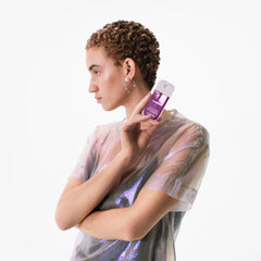 Woman with short curly hair in an iridescent sheer top holding Cashmere Woods Power Essence fragrance mist