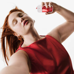 Red-haired woman in a red satin top holding Golden Amber Power Essence body and hair mist overhead