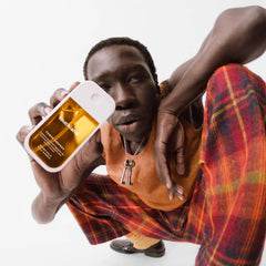 Man in plaid trousers and an orange knit vest holding up Mango Mojo Power Essence body and hair mist