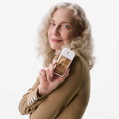 Woman with long silver curly hair in a camel blouse holding Vanilla Velvet Power Essence fragrance mist at her shoulder