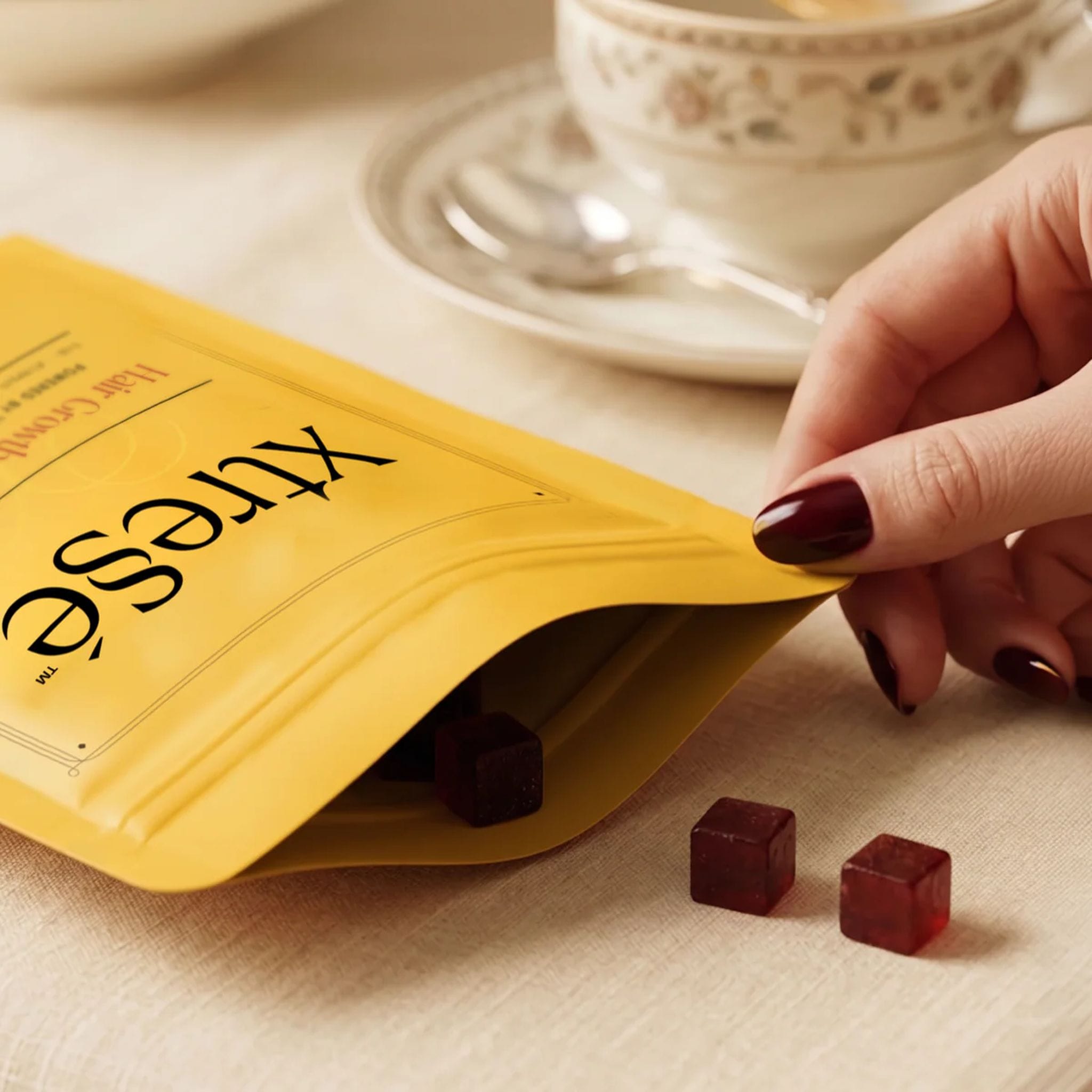 Hand opening Xtressé hair growth gummies pouch beside a teacup