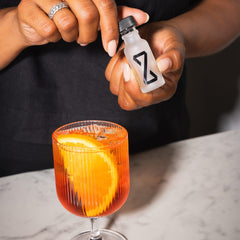 Hands opening ZBiotics Pre-Alcohol Probiotic Drink bottle above orange cocktail with citrus garnish on marble bar