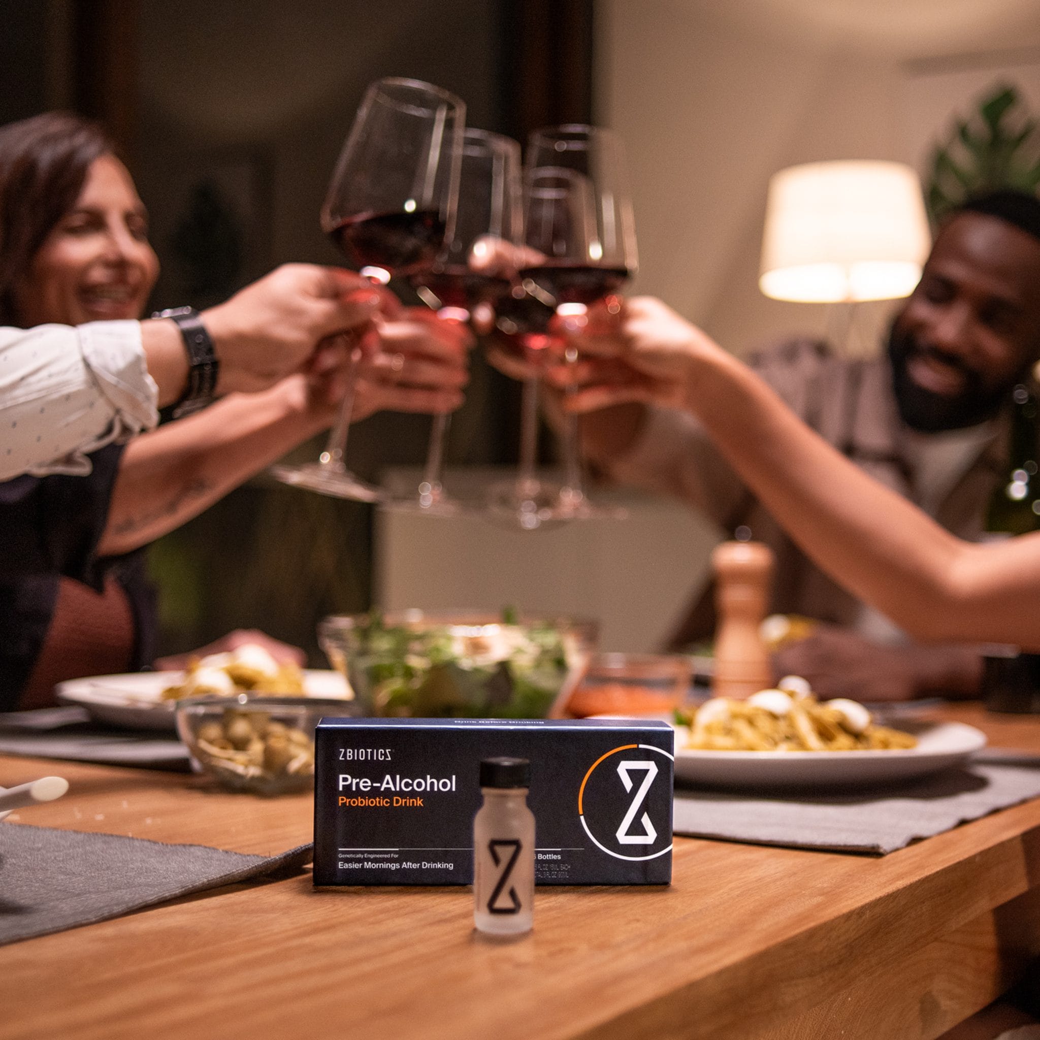 Friends toasting wine glasses at dinner table with ZBiotics Pre-Alcohol Probiotic Drink box and bottle