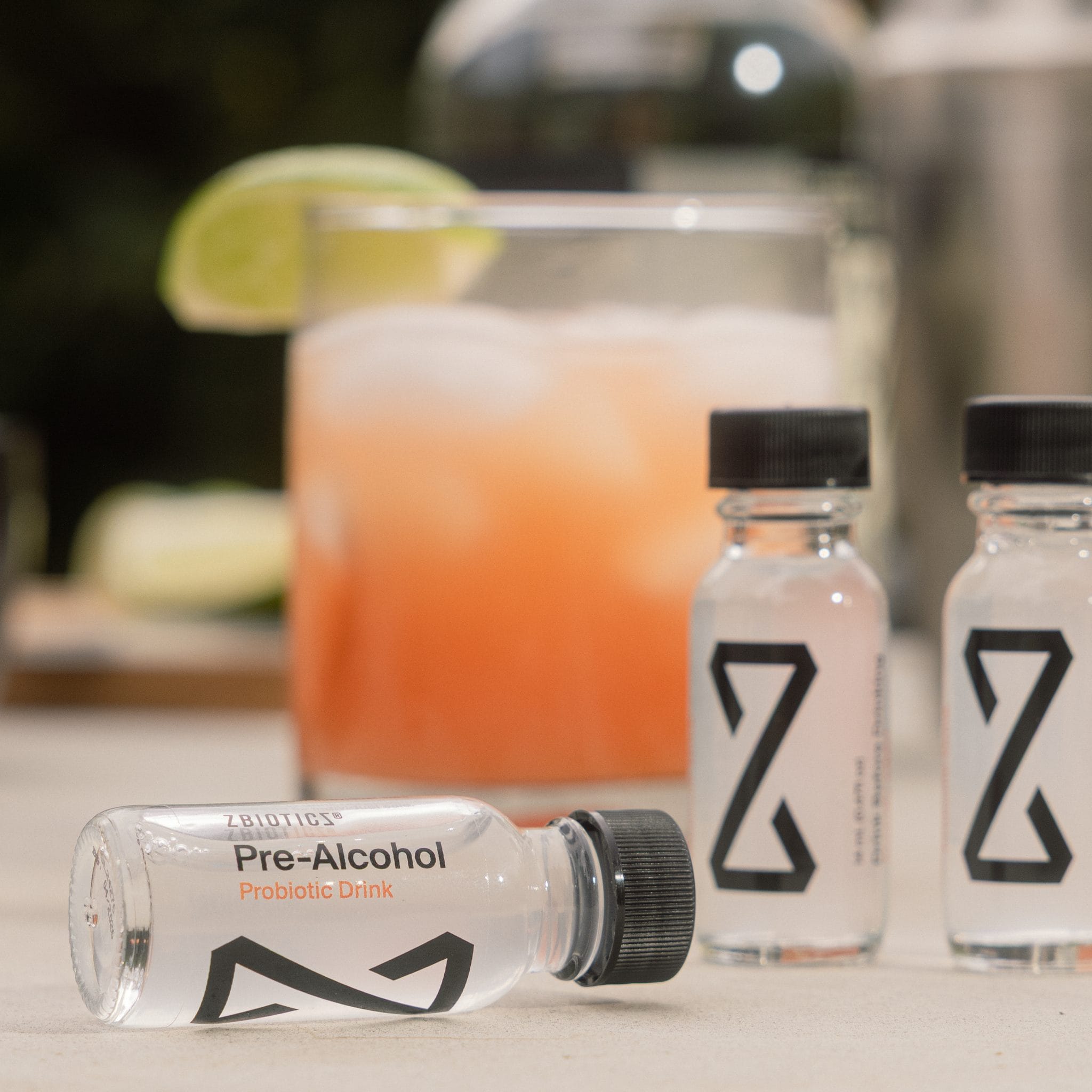 Three ZBiotics Pre-Alcohol Probiotic Drink bottles with pink cocktail and lime garnish outdoors